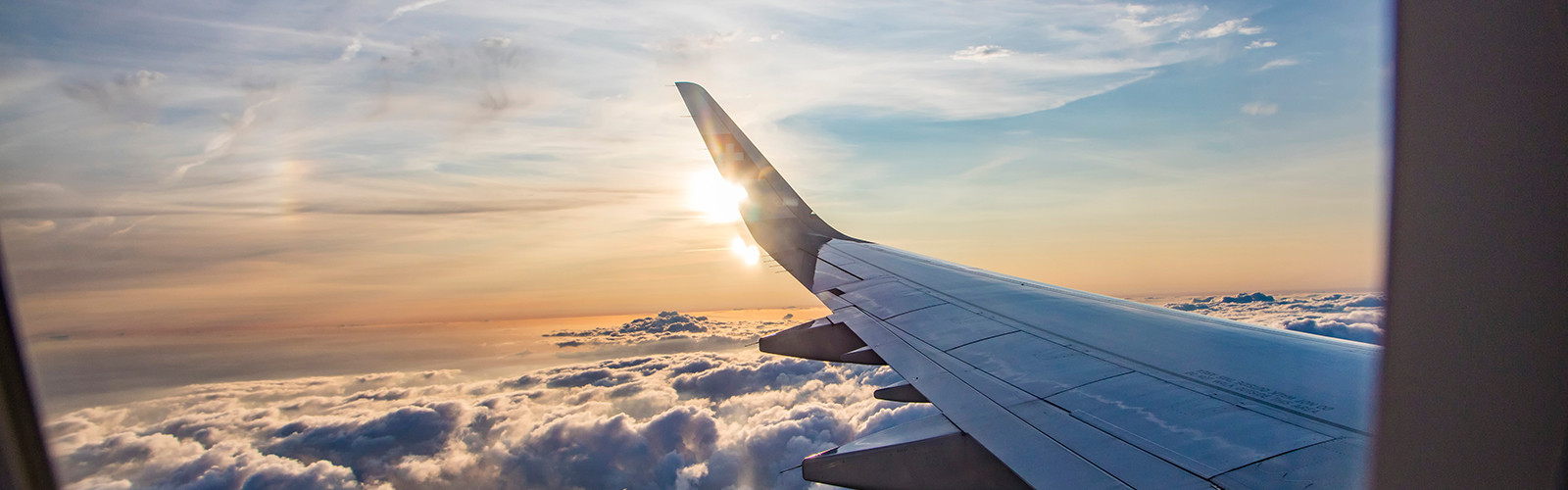 Airplane wing above clouds during sunrise, sunlight reflecting on metal.