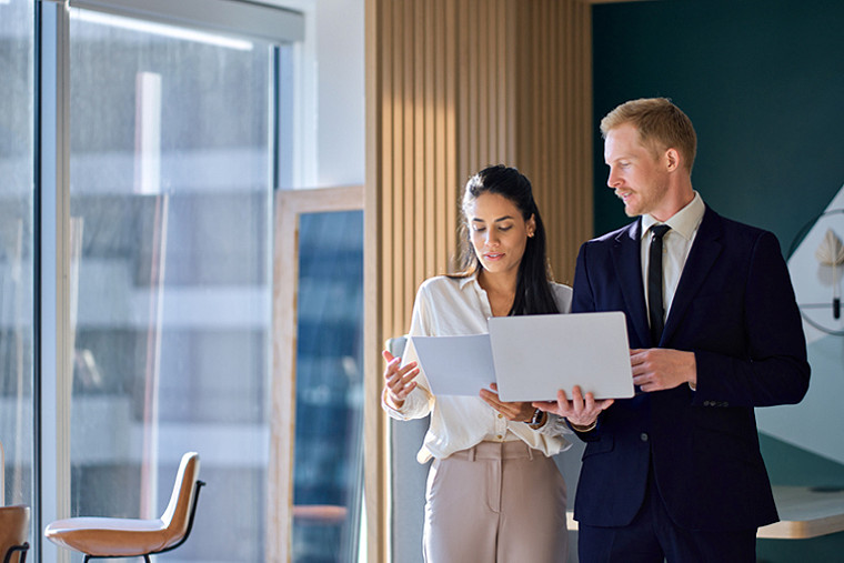 Two business people standing in front of a large window, discussing work.