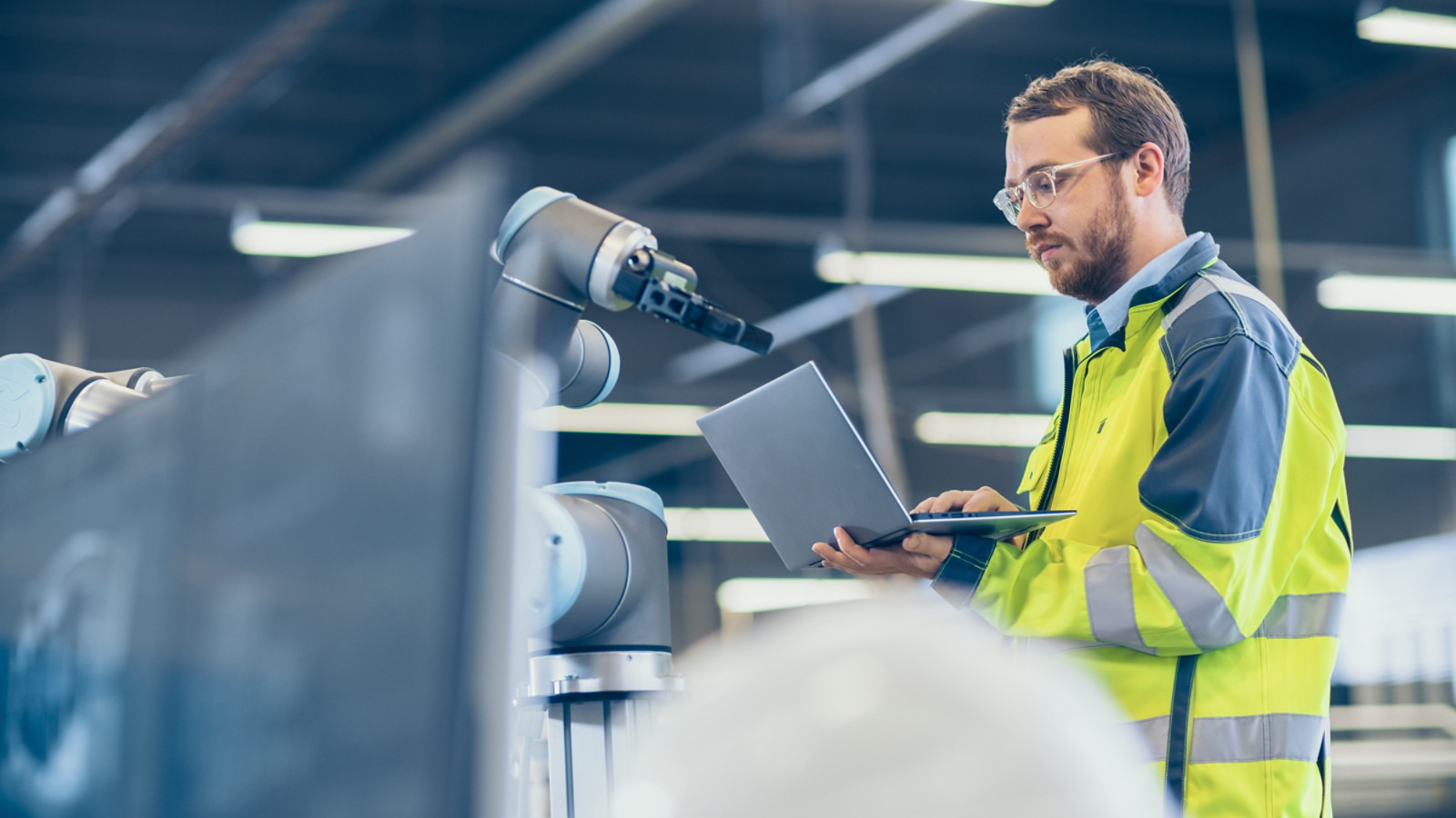 At the Factory: Automation Engineer Uses Laptop for Programming Robotic Arm. New Era in Automatic Manufacturing Industry.