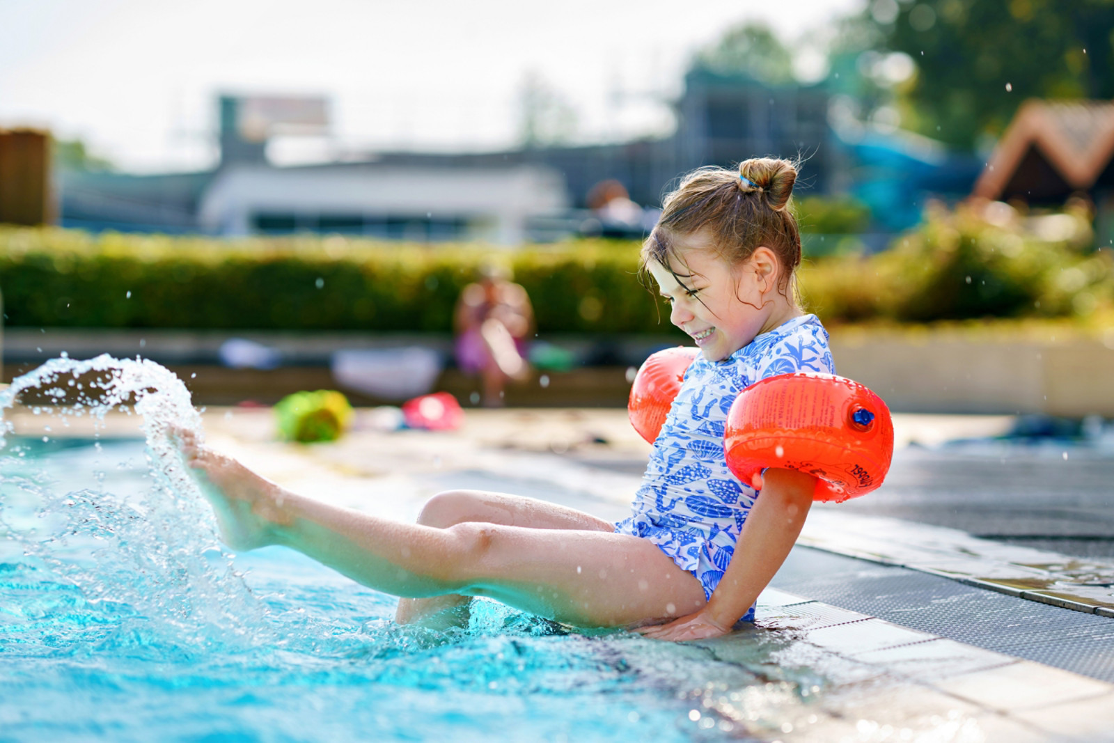 Little preschool girl with protective swimmies playing in outdoor swimming pool by sunset. Child learning to swim in outdoor pool, splashing with water, laughing and having fun. Family vacations