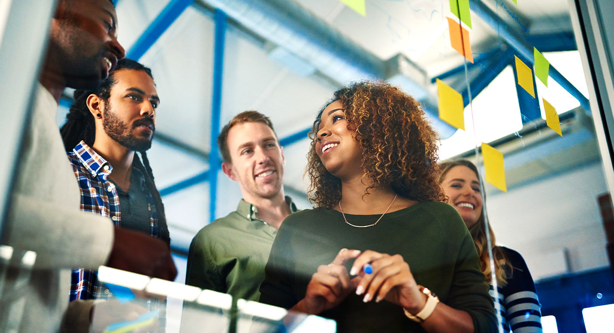A diverse group of five people in an office, smiling and brainstorming around a glass board with sticky notes. The atmosphere is collaborative and creative.