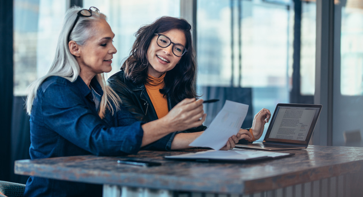 Dos mujeres sentadas en una mesa, que trabajan en un portátil, hablando