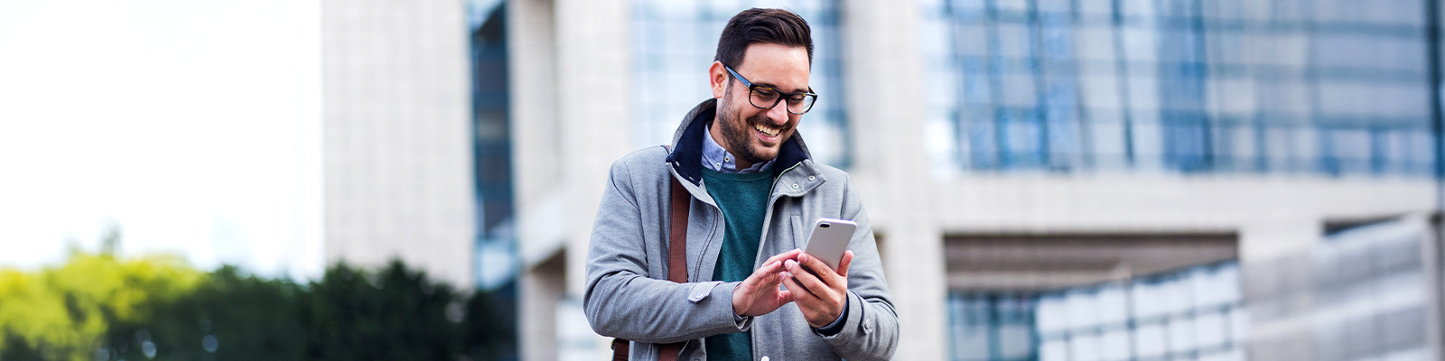A man in a gray jacket and glasses smiles while using a smartphone outdoors. He stands in front of a modern glass building, conveying a cheerful mood.