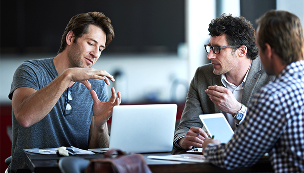 Three men engaged in discussion around a table with a laptop and tablet, looking focused and collaborative in a modern office setting.