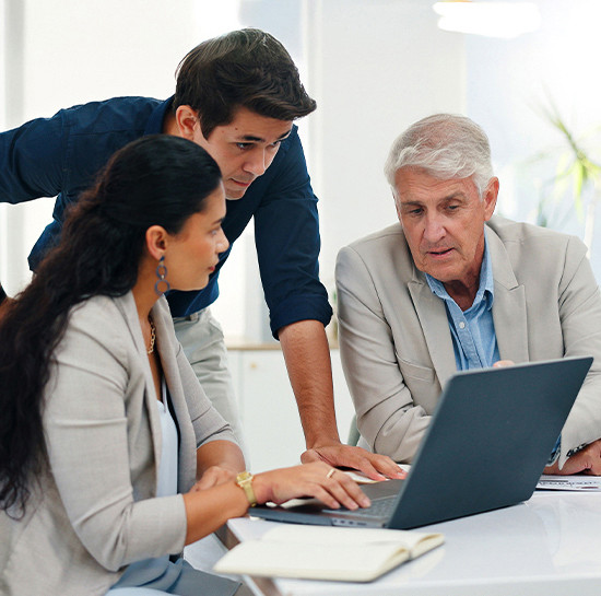 Three people in a focused discussion around a laptop in a bright office. Two men and a woman collaborate, suggesting teamwork and planning.