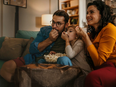 A family of three sitting on a couch, munching on popcorn and, possibily, watching television.