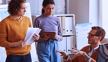 A couple of young employees standing and listening to a senior person sitting and talking to them
