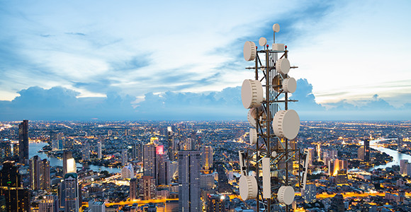 A tall telecommunications tower with numerous antennae overlooks a sprawling cityscape at dusk. The sky is clear, with a soft blue hue and scattered clouds.