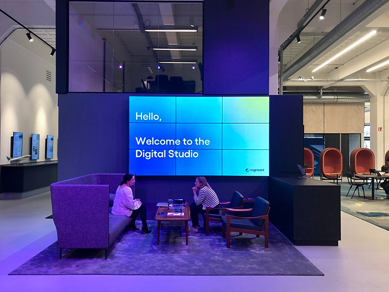 Two women sitting across a coffee table at Cognizant's Amsterdam office, discussing, with a big digital display on the wall