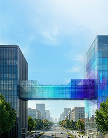 A transparent fibreglass bridge connecting two tall glass-faced buildings over a busy street in a city
