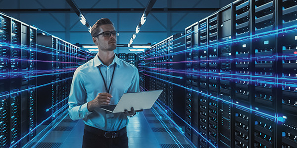 In a room with servers stacked on either side, a man holding an open laptop is standing and looking at something.