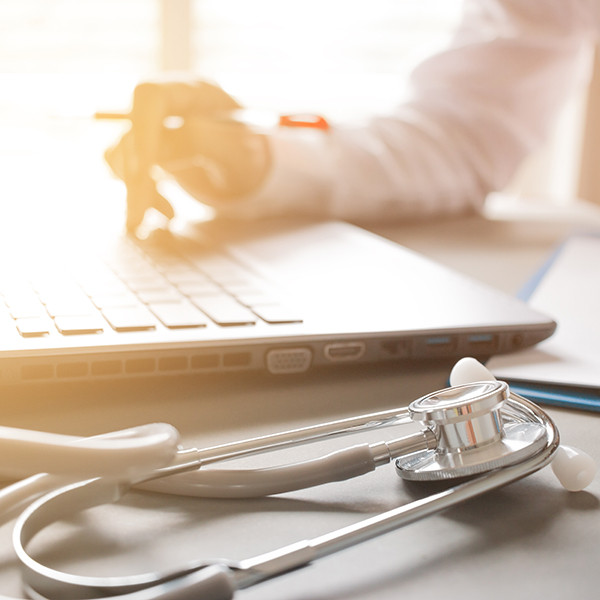 Hand holding a pen hovering over keyboard and a stethoscope lying beside the laptop