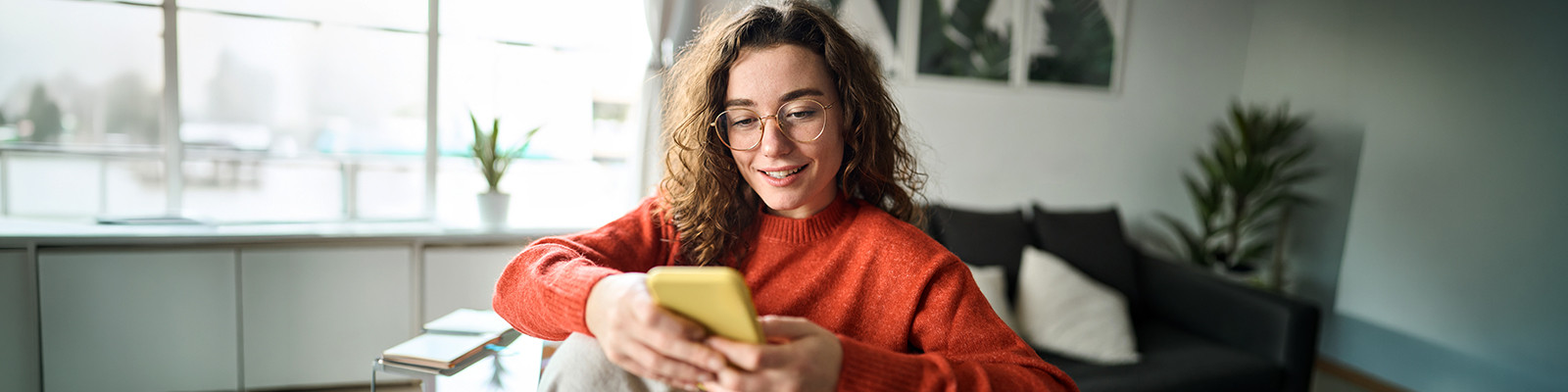 Person using a smartphone while relaxing in a bright, modern living space with natural light, comfortable furnishings, and indoor plants.