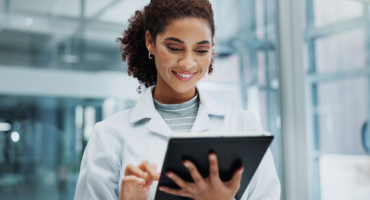 A woman in a lab coat smiles while using a tablet, standing in a bright, modern office with large glass windows