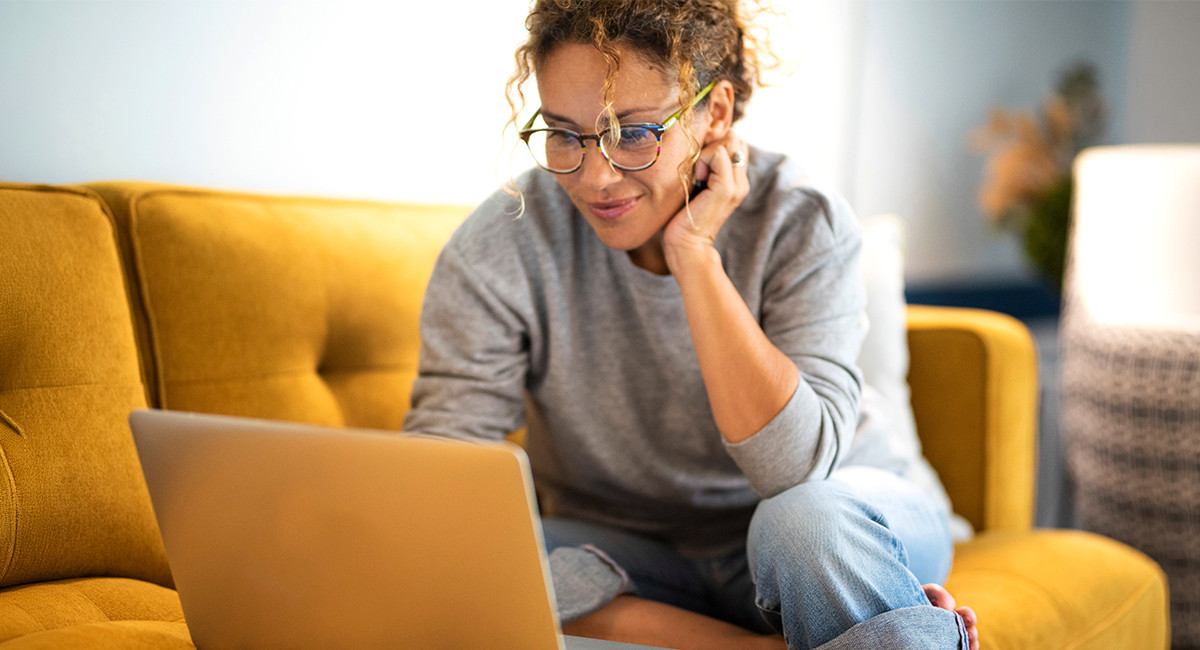 Person wearing glasses uses a laptop while sitting on a yellow sofa in a bright home setting.