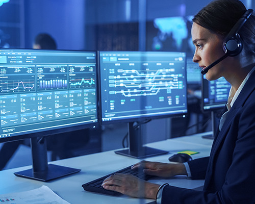 Mujer con auriculares, sentada frente a varios monitores y escribiendo en un teclado