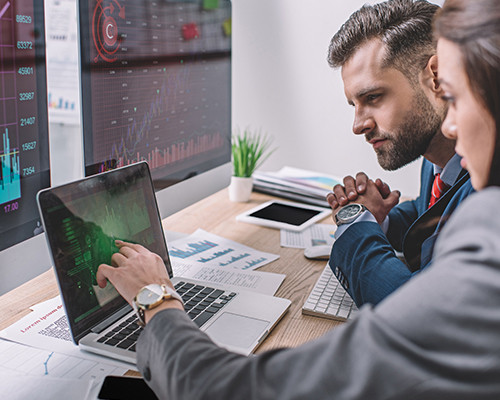 Ein Mann und eine Frau mit Profil­ansicht sitzen an einem Schreibtisch und schauen auf einen Laptop