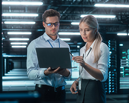 Woman and man standing in a server room, facing us, looking at a laptop