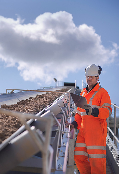 A worker wearing an orange overall standing, standing outdoors next to a conveyor belt, looking at a tablet