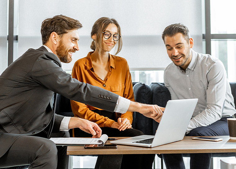 A 3-people discussion at an office table with one man pointing to a laptop screen