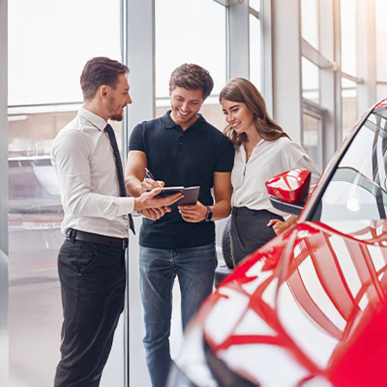A male and female couple and a salesman looking at a tablet while standing by a new car.