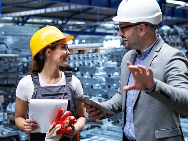 Agirl wearing a yellow helmet and gray overalls holding a clipboard listening to a man in a gray suit and white helmet, holding a tablet and talking