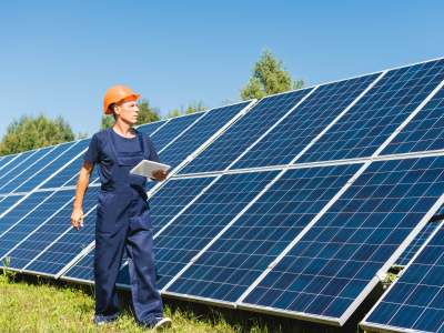A man in a blue overall and orange helmet holding a tablet and walking in front of a row of solar panels