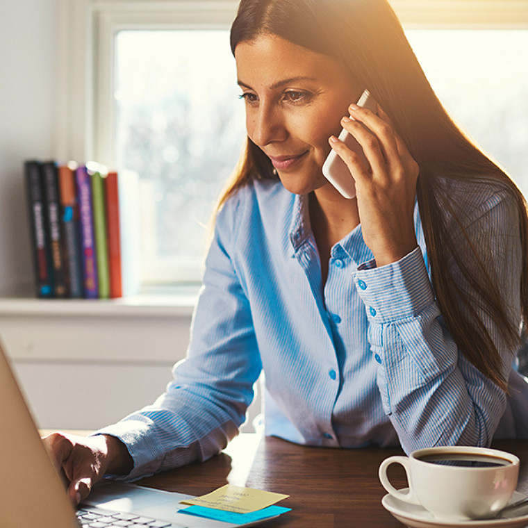 A woman, backlit in warm light, working on laptop while talking on phone