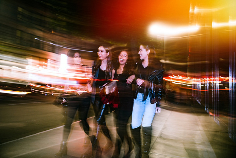 A group of four girlfriends walking arm-in-arm along  a busy, well lit street, happily laughing and talking