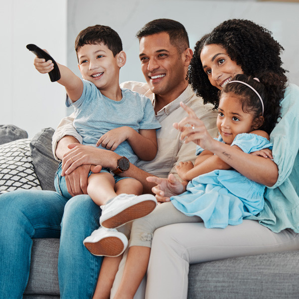 A family kind of picture with a man, woman and two kindssitting on a sofa and watching television with the remote on the hand