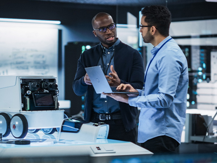 Two male colleagues standing, one talking while the other listens, holding an open laptop