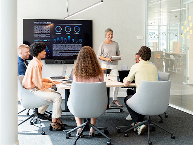 Five people sitting around a table, while a lady is standing and making a presentation
