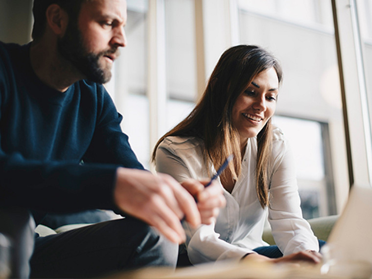 A woman is sitting and working on a laptop, while her partner looks on