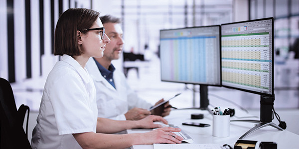 A couple of professionals wearing white lab jackets, sitting at a desk and looking at data on a computer screen.