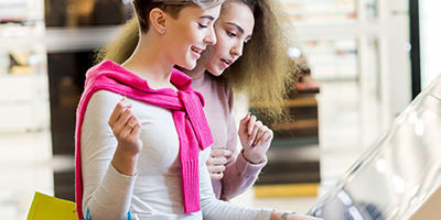 Two adult caucasian girlfriends use information kiosk at shopping center. Happy women with shopping bags pointing finger at the info desk. Consumerism, shopping, lifestyle concept