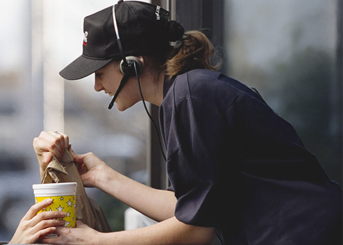 A fast-food drive-through employee wearing a headset is hanging out of the window and handing out a packet and drink to a customer