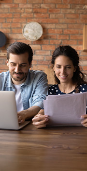 Ein Mann und eine Frau unterhalten sich lächelnd, sitzen dabei an einem Tisch und schauen sich Dokumente und einen Laptop an
