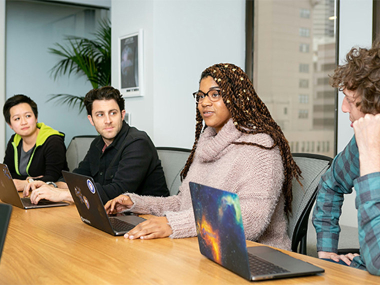 Four people seated at a meeting with laptops open in front of them on the table