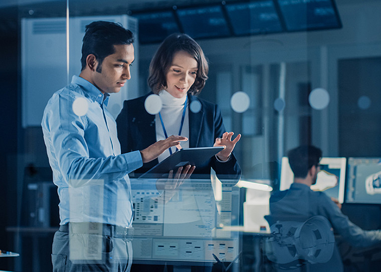 Two colleagues in an office discussing data on a tablet, with digital dashboard overlays and a colleague working at monitors in the background, conveying collaborative tech innovation.