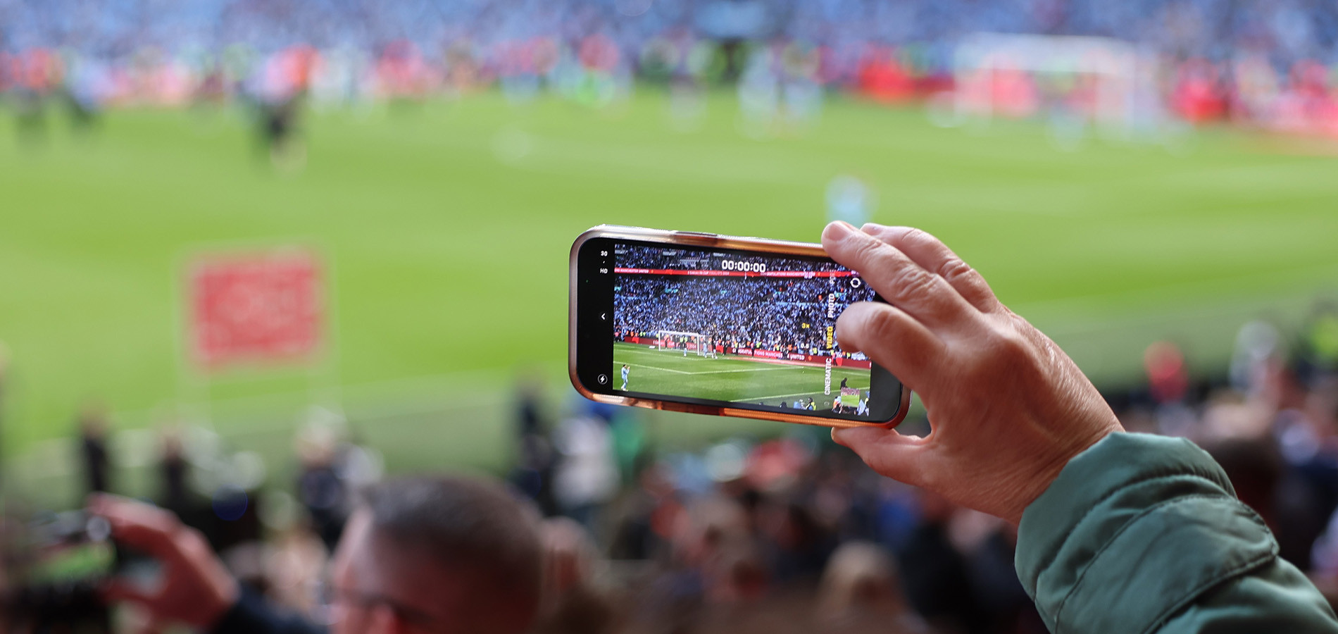 A hand holding a mobile phone, photographing the soccer stadium thatis in the background