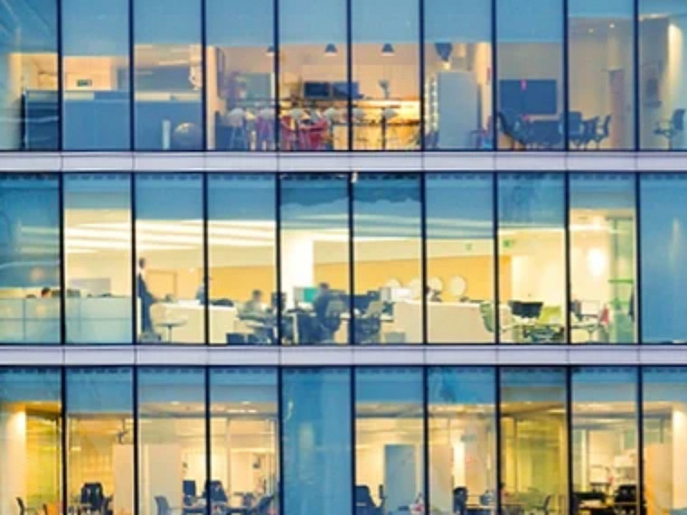 Office building windows showing multiple brightly lit workspaces inside