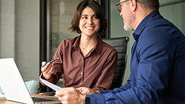 Two colleagues in business attire share a discussion at a desk with a laptop. The woman smiles, creating a positive and collaborative atmosphere.