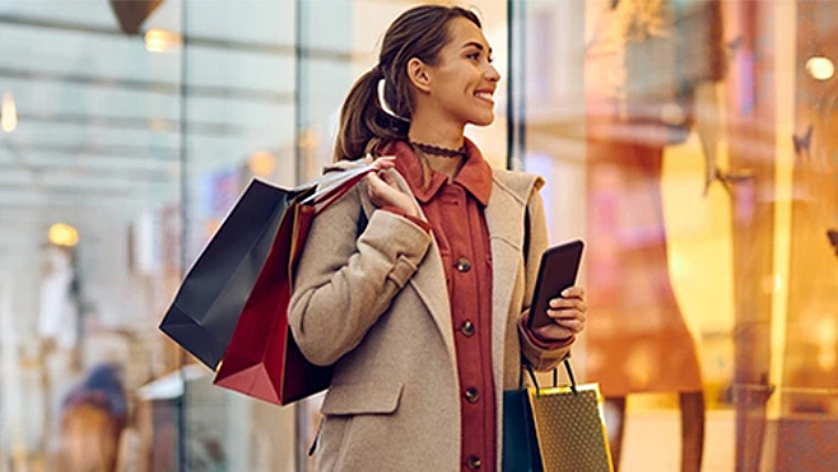 A woman carrying several shopping bags, is smiling and looking at products displayed in a store window