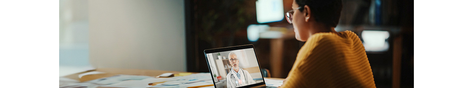 A person wearing glasses and sitting at a table is having a video call with a doctor displayed on a laptop screen.