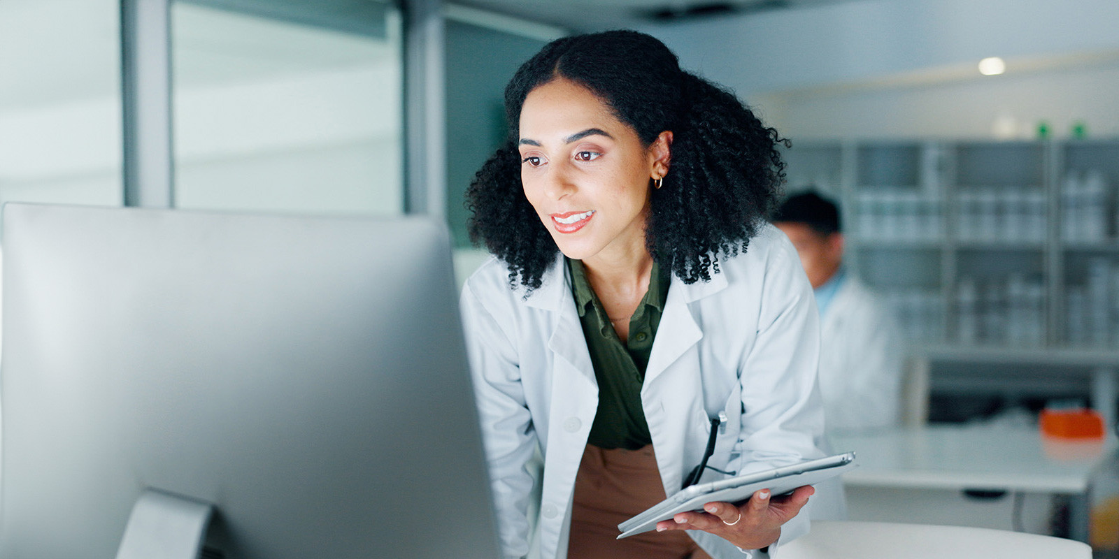 Healthcare professional in a clinical lab reviewing data on a desktop computer while holding a tablet.