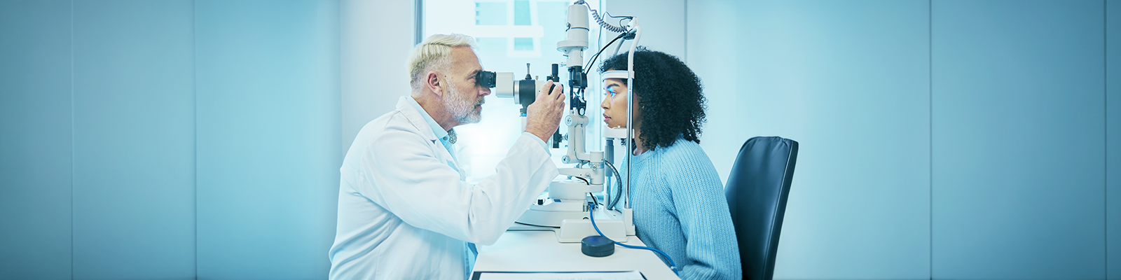 An optometrist examines a patient's eyes using diagnostic equipment in a brightly lit clinic.