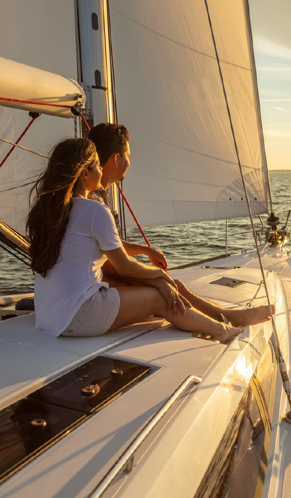 Man and woman sitting on sailboat looking out to sun over body of water