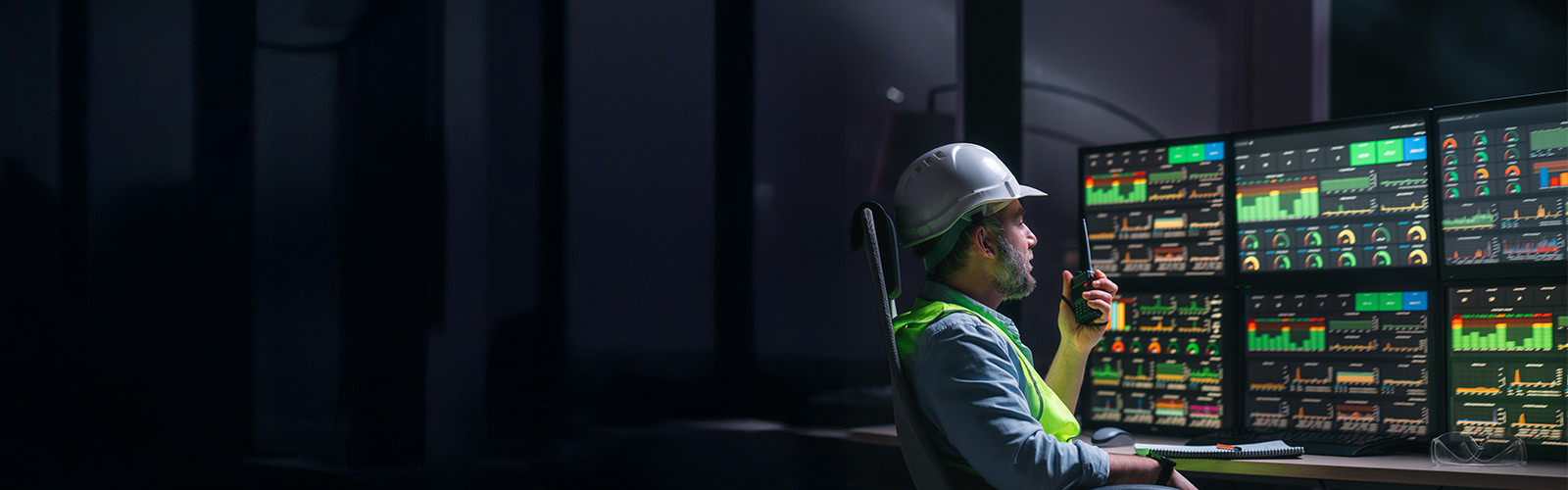 A male worker in hard hat and safety vest sitting in front of multiple computer monitors.