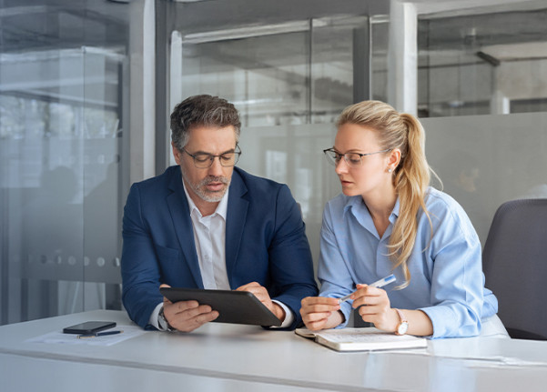A man and a woman in business attire sit at a table, focused on a tablet. The setting is an office with a calm, professional atmosphere.