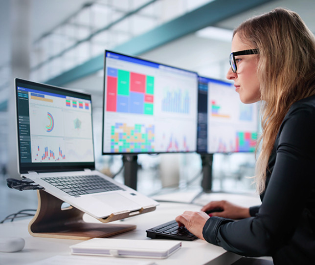 Woman in an office studies data analytics on three monitors, focusing intently. The setting is modern and professional, conveying concentration.